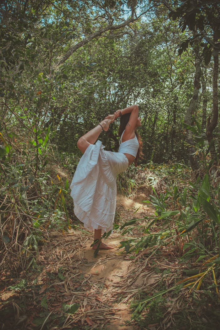 A woman in a white dress performs a yoga pose surrounded by lush forest greenery.